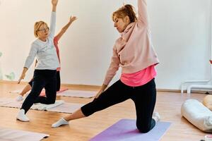 A group of senior women engage in various yoga exercises, including neck, back, and leg stretches, under the guidance of a trainer in a sunlit space, promoting well-being and harmony photo