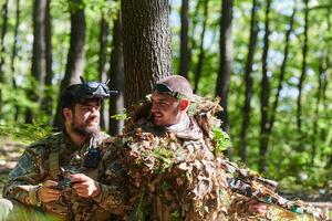 A skilled sniper and a soldier operating a drone with VR goggles strategize and observe the military action while concealed in the forest photo