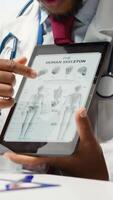 Vertical African american specialist showing the human skeleton to explain diagnosis to patient, x ray imaging scan interpretation. Medical expert reviewing lab test results in cabinet. Camera A. video