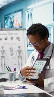 Vertical African american specialist showing the human skeleton to explain diagnosis to patient, x ray imaging scan interpretation. Medical expert reviewing lab test results in cabinet. Camera B. video