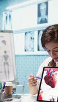 Vertical Woman specialist explaining heart function and condition on test results, doctor patient interaction in a cabinet. Physician cardiologist showing the cardiovascular system on tablet. Camera B. video