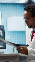 Vertical Female patient listening to a diagnostic report from doctor based on MRI scan results and x ray imaging, pointing at computer in medical cabinet. African american expert gives assistance. Camera A. video