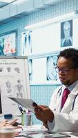 Vertical Female patient listening to a diagnostic report from doctor based on MRI scan results and x ray imaging, pointing at computer in medical cabinet. African american expert gives assistance. Camera B. video