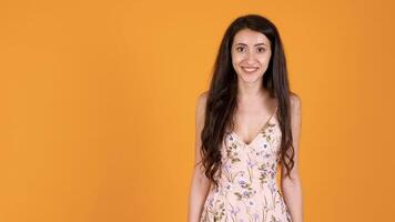 Gorgeous young woman in summer dress looking surprised at camera isolated on yellow orange background in studio video
