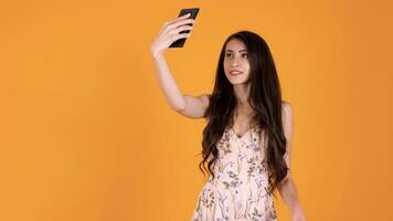 Gorgeous young woman in summer dress taking a selfie isolated on yellow orange background in studio video