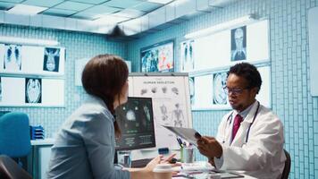 Female patient listening to a diagnostic report from doctor based on MRI scan results and x ray imaging, pointing at computer in medical cabinet. African american expert gives assistance. Camera B. video