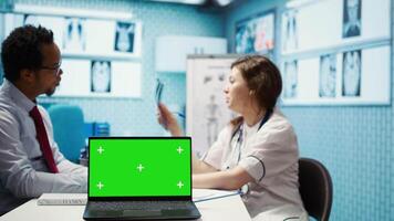 General practitioner and patient looking at green screen on device, recommending pills and drugs during check up visit. Diverse people discussing about diagnostics and remedy. Camera B. video
