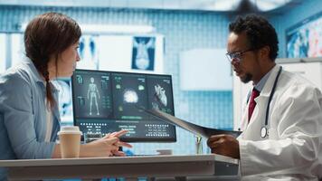 Female patient listening to a diagnostic report from doctor based on MRI scan results and x ray imaging, pointing at computer in medical cabinet. African american expert gives assistance. Camera A. video