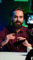 Vertical Upbeat content creator hosting online show in rgb lights studio, shooting vlog using professional microphone. Cheerful vlogger presenting informations during streaming service broadcast, camera A video