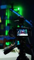 Vertical Focus on professional camera used by man in blurry background hosting online show in neon lit studio. Close up of high tech gear used by vlogger during streaming service broadcast, camera B video