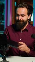 Vertical Man in studio endorsing laptop from brand sponsoring , filming with smartphone on tripod. Tech expert does influencer marketing by promoting notebook, recording with phone, camera A video