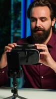 Vertical Media star in neon lit studio presents VR goggles from brand sponsoring , closely analyzing features. Influencer reviewing virtual reality headset for tech enthusiasts watching, camera A video