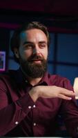 Vertical Happy influencer in home studio greeting audience, recording live broadcast using smartphone on selfie stick, close up. Man filming q and a session with mobile phone, answering questions, camera B video