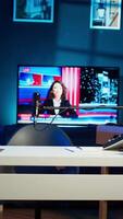 Vertical Work desk with laptop and house plants in empty house interior. Living room illuminated by neon lights with notebook device sitting on table, news broadcast running on TV in background video