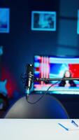 Vertical TV screen showing news broadcast and recording mic in empty stylish neon lit home studio interior. Streaming microphone in modern apartment illuminated with RGB lights at night and television set turned on video