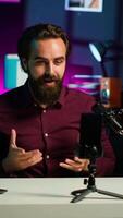 Vertical Media star talking in front on smartphone attached to tripod stabilizer in studio, discussing with viewers. Man using mobile phone to record in living room for social media, camera A video