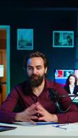 Vertical Joyful content creator sitting at table talking in microphone in dimly lit home studio interior, chatting with subscribers. Upbeat man surrounded by podcast recording gear in apartment, camera B video