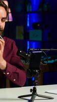 Vertical Tech guru presenting V mount battery, filming technology review using cellphone on tripod and professional microphone. Caucasian vlogger showcases charging device specs, recording with phone, camera A video