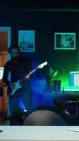 Vertical Playful internet star playing guitar in studio while shooting . Energetic content creator playing musical instrument while filming vlog with professional mic in home studio, camera B video