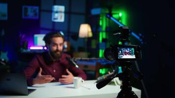 Focus on professional camera used by man in blurry background hosting online show in neon lit studio. Close up of high tech gear used by vlogger during streaming service broadcast, camera B video