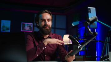 Happy influencer in home studio greeting audience, recording live broadcast using smartphone on selfie stick, close up. Man filming q and a session with mobile phone, answering questions, camera B video