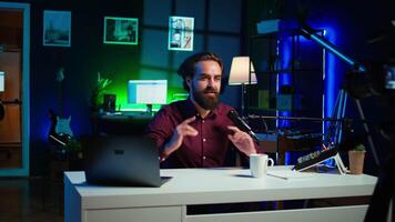 Upbeat content creator hosting online show in rgb lights studio, shooting vlog using professional microphone. Cheerful vlogger presenting informations during streaming service broadcast, camera B video