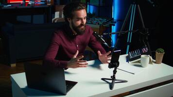 Media star talking in front on smartphone attached to tripod stabilizer in studio, discussing with viewers. Man using mobile phone to record in living room for social media, camera B video
