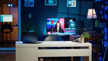 Work desk with laptop and house plants in empty house interior. Living room illuminated by neon lights with notebook device sitting on table, news broadcast running on TV in background video