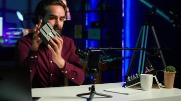 Tech guru presenting V mount battery, filming technology review using cellphone on tripod and professional microphone. Caucasian vlogger showcases charging device specs, recording with phone, camera A video