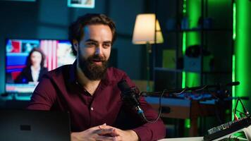 Joyful content creator sitting at table talking in microphone in dimly lit home studio interior, chatting with subscribers. Upbeat man surrounded by podcast recording gear in apartment, camera A video