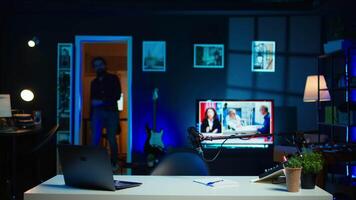 Happy influencer using microphone, arriving in dimly lit home studio interior, chatting with fans. Smiling man surrounded by podcast recording equipment in apartment starting stream, camera B video