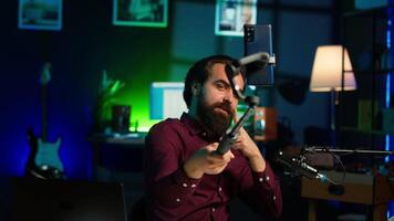 Influencer doing peace sign in front on cellphone attached to selfie stick in studio, chatting with viewers using mic. Man doing hand gesturing, using phone and microphone to record vlog, camera A video