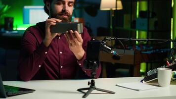 Influencer does technology review of wireless portable speaker for internet platforms channel, filming with mobile phone. Influencer showing music playing device, recording with smartphone, camera A video