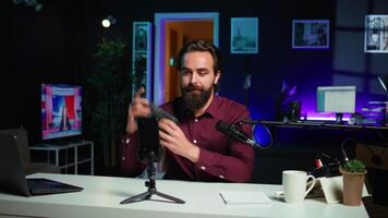 Tech guru presenting V mount battery, filming technology review using cellphone on tripod and professional microphone. Caucasian vlogger showcases charging device specs, recording with phone, camera B video