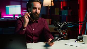Content creator talking in front on cellphone attached to selfie stick in studio, chatting with viewers using recording mic. Man using phone camera and professional microphone to record vlog, camera A video