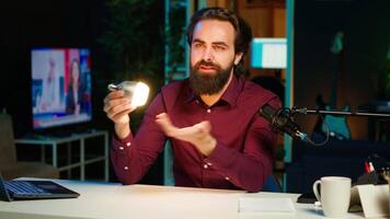Tech guru in dimly lit apartment studio filming smart light bulb review for online streaming platforms. Media star hosting show, presenting IoT home automation product, camera A video