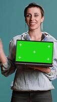 Vertical Young woman pointing at laptop with green screen display in studio, using innovative technology on wireless pc. Cool fashionable person does a bold advertisement. Camera B. video
