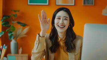 Smiling Asian Woman Waving During Vibrant Call in Modern Office Setting photo