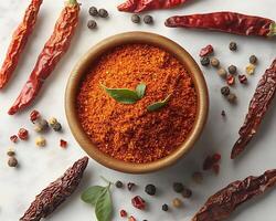 Small bowl of freshly ground cayenne pepper placed in the middle of a white background surrounded by whole dried chilies photo