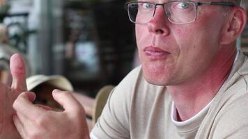 Young blond man in glasses sits at a table in a cafe, wipes his hands with a napkin after eating and blows his nose into a handkerchief, Blurred background, rest after a hearty lunch, video