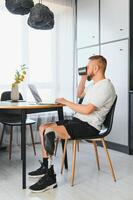 Man amputee with prosthetic leg disability on above knee transfemoral leg prosthesis artificial device using laptop at home. People with amputation disabilities everyday life photo