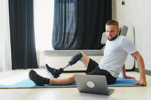 Sporty fit young man amputee with prosthetic leg disability prosthesis doing training exercises on mat at home using laptop. Inclusive online sport classes for people with disabilities concept photo