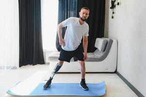 Man amputee with prosthetic leg disability on transfemoral leg prosthesis artificial device making stretching rehabilitation physical exercise at home. People w amputation disabilities everyday life photo