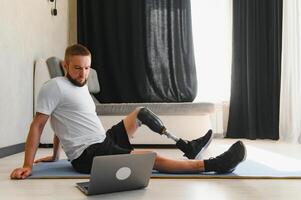 Sporty fit young man amputee with prosthetic leg disability prosthesis doing training exercises on mat at home using laptop. Inclusive online sport classes for people with disabilities concept photo