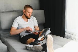 Man amputee with prosthetic leg disability on above knee transfemoral leg prosthesis artificial device using laptop at home. People with amputation disabilities everyday life photo