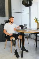 Man amputee with prosthetic leg disability on above knee transfemoral leg prosthesis artificial device using laptop at home. People with amputation disabilities everyday life photo