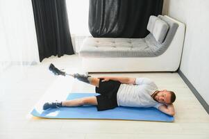 Full length portrait of man with prosthetic leg doing stretching exercises at home and reaching toes photo