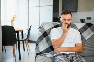 Dental health problems concept. Man suffering from strong toothache. Upset male patient with inflamed nerve, tooth abscess, or teeth sensitivity sitting on sofa and touching cheek with grimace of pain photo