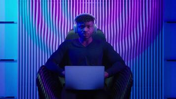 African American man guy male coder gamer programmer in chair wear headphones using laptop online work crypto software play internet game modern technology neon futurism showing thumb up like gesture video