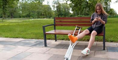 Teenage girl with a leg in a cast seated on a park bench, utilizing her smartphone to browse social media and content while surrounded by the tranquil outdoor environment photo